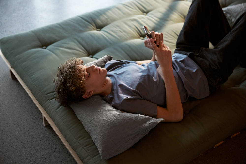 En ung kille ligger på soffan och tittar på sin mobil.